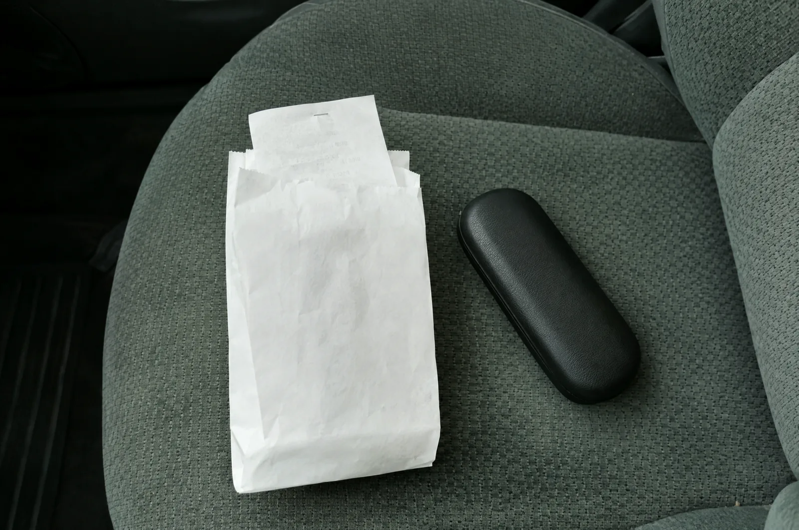 Overhead of a car's passenger seat with a plain white pharmacy bag, a receipt half out of it and a dark glasses case beside it.