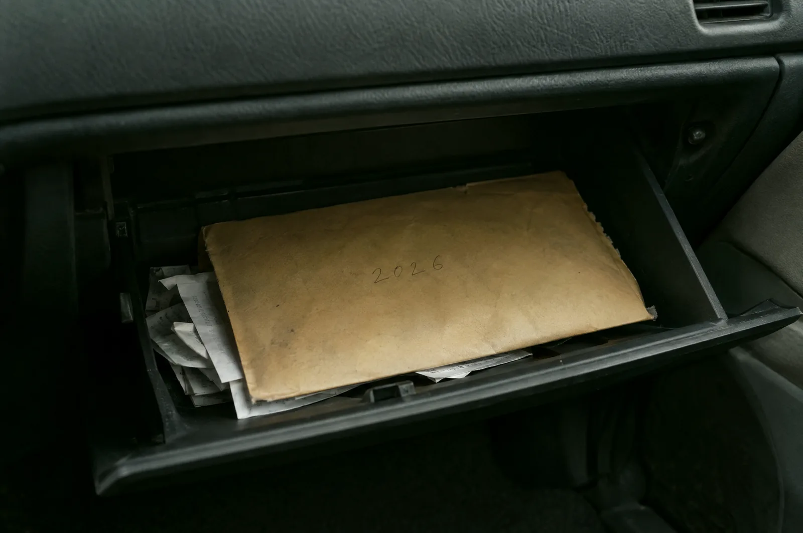 An open glovebox holding a worn manila envelope marked 2026 in pencil, with a few folded receipts inside.