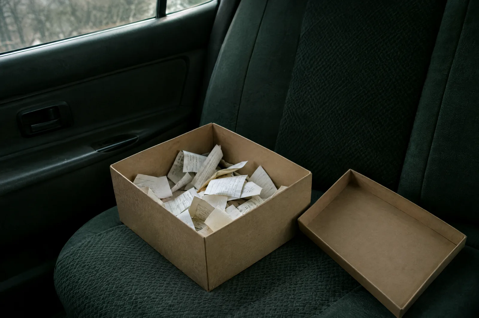 A cardboard shoebox with its lid set aside on a car's back seat, filled with a loose jumble of curled paper receipts.
