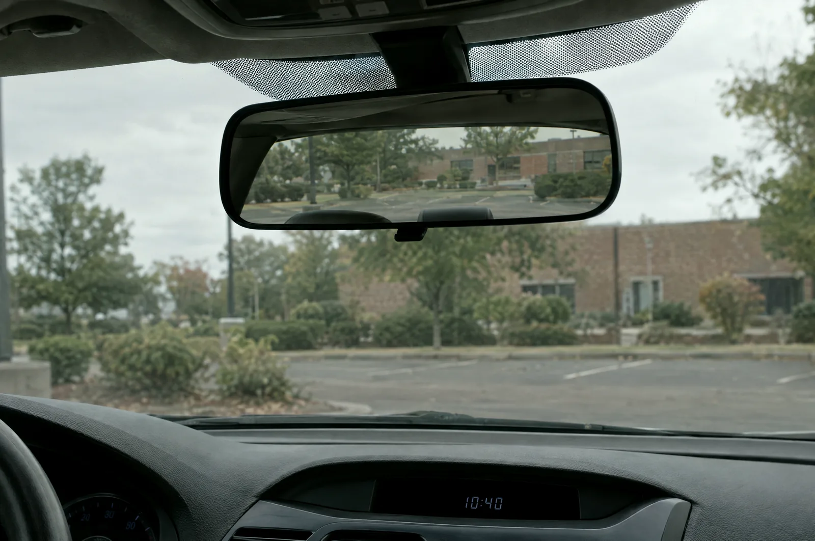 A car's rearview mirror and upper windshield with a parking lot and a low brick building reflected, the dashboard clock reading 10:40.